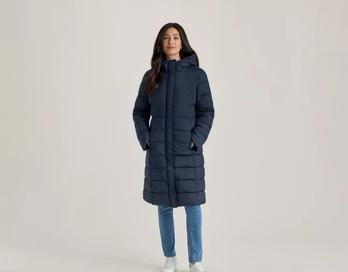 Frau steht, trägt einen langen, marineblauen, wattierten Mantel mit Kapuze, hellblaue Jeans und weiße Turnschuhe, die Hände in den Taschen.