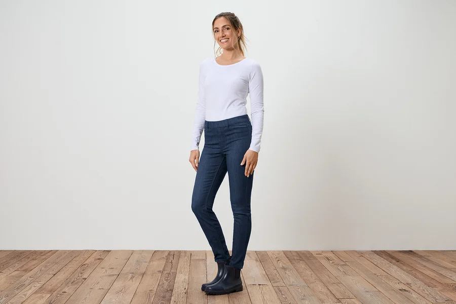 Eine Frau steht seitlich in Jeans, einem weißen Langarmshirt und Gummistiefeln vor einer weißen Wand und einem Holzboden.
