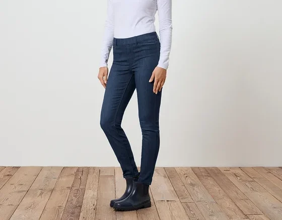 Eine Frau steht seitlich in Jeans, einem weißen Langarmshirt und Gummistiefeln vor einer weißen Wand und einem Holzboden.