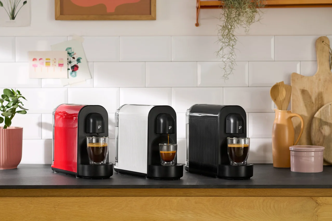 Drei Kaffeemaschinen in Rot, Weiß und Schwarz stehen nebeneinander auf einer Küchentheke, wobei jede Maschine Kaffee in eine darunter stehende Tasse brüht.
