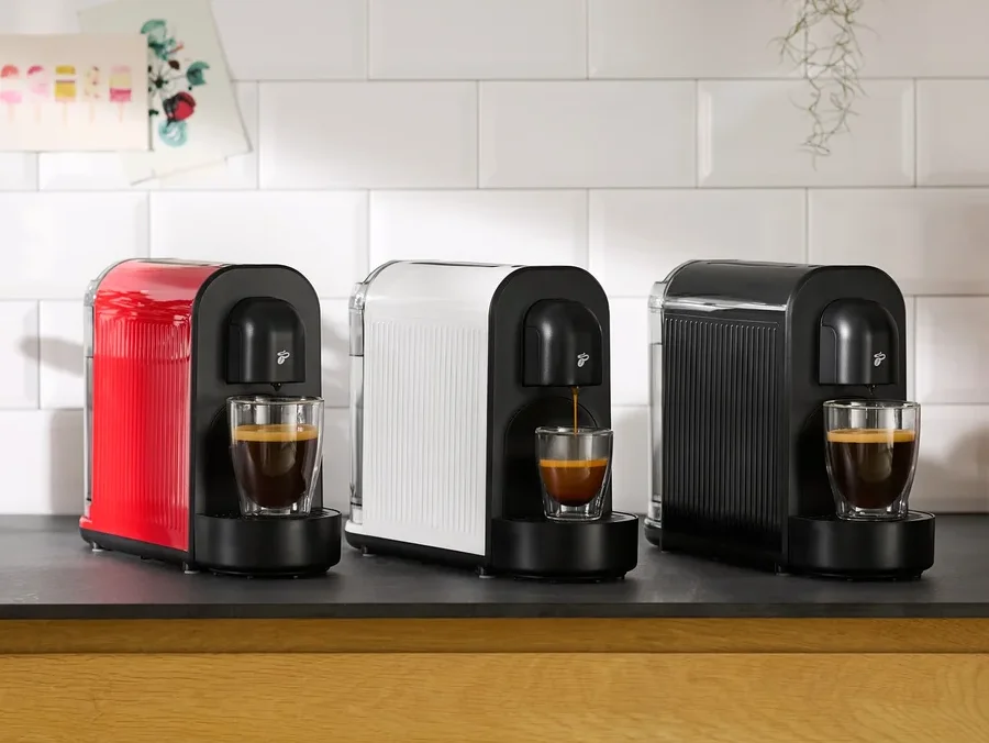 Drei Kaffeemaschinen in Rot, Weiß und Schwarz stehen nebeneinander auf einer Küchentheke, wobei jede Maschine Kaffee in eine darunter stehende Tasse brüht.
