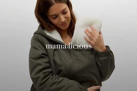 Eine lächelnde Frau mit braunen Haaren trägt eine grüne Jacke mit weißem Teddyfutter und hält ein Baby oder einen Trageeinsatz mit weißem Fell an ihrer Brust.