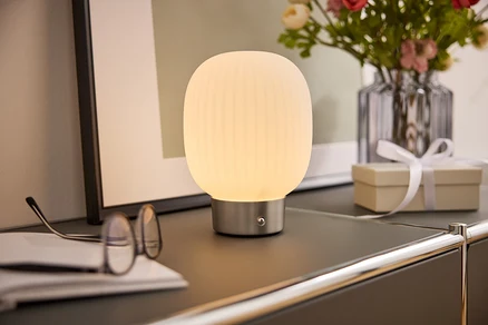 Eine Nachttischlampe mit weißem Schirm steht auf einem Schrank neben einer Brille, einem Buch, einer Blumenvase und einer Geschenkbox.