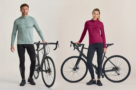Ein Mann und eine Frau stehen neben schwarzen Rennrädern. Der Mann trägt ein hellgrünes langärmeliges Radtrikot und eine schwarze Radhose. Die Frau trägt ein dunkelrosa langärmeliges Radtrikot und eine schwarze Radhose.