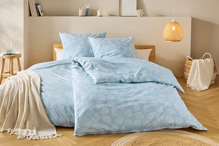 Ein Schlafzimmer zeigt ein hellblaues Bettzeug mit Muschelmuster, einen Holzhocker mit Decke und eine geflochtene Lampe.