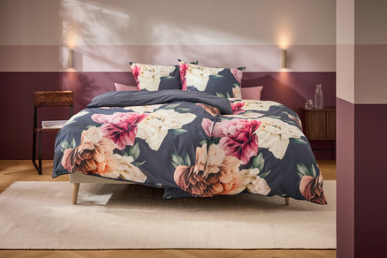 Ein Bett mit dunkelgrauer Bettwäsche und großen rosa, weißen und orangefarbenen Blumenmustern steht in einem Schlafzimmer mit zweifarbiger Wand und Holzmöbeln.
