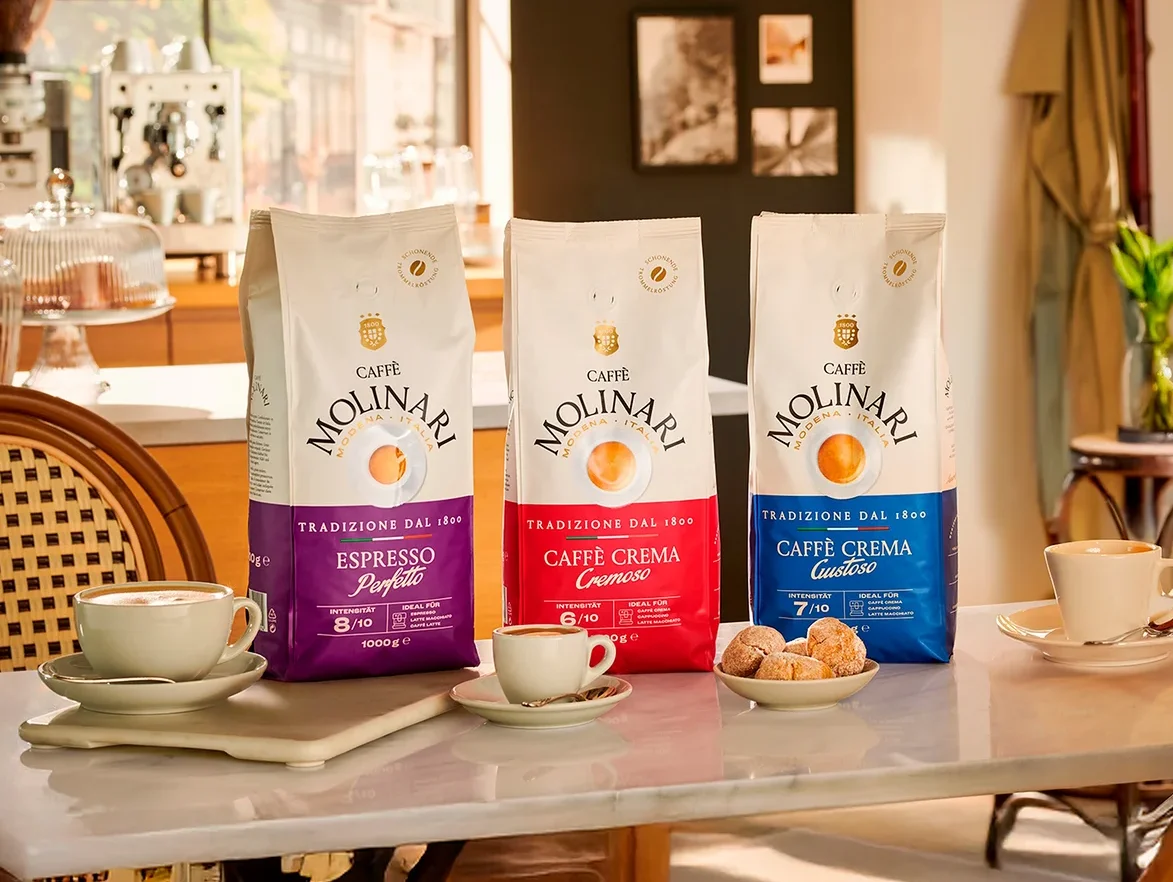 Drei Packungen Molinari Kaffee stehen auf einem Tisch neben Tassen und Tellern mit Gebäck.