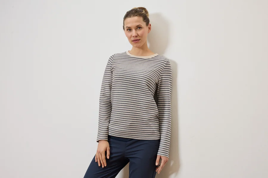 Frau mit Dutt trägt ein grau-weiß gestreiftes Langarmshirt und eine dunkelblaue Hose vor einer weißen Wand.