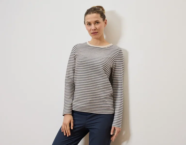 Frau mit Dutt trägt ein grau-weiß gestreiftes Langarmshirt und eine dunkelblaue Hose vor einer weißen Wand.
