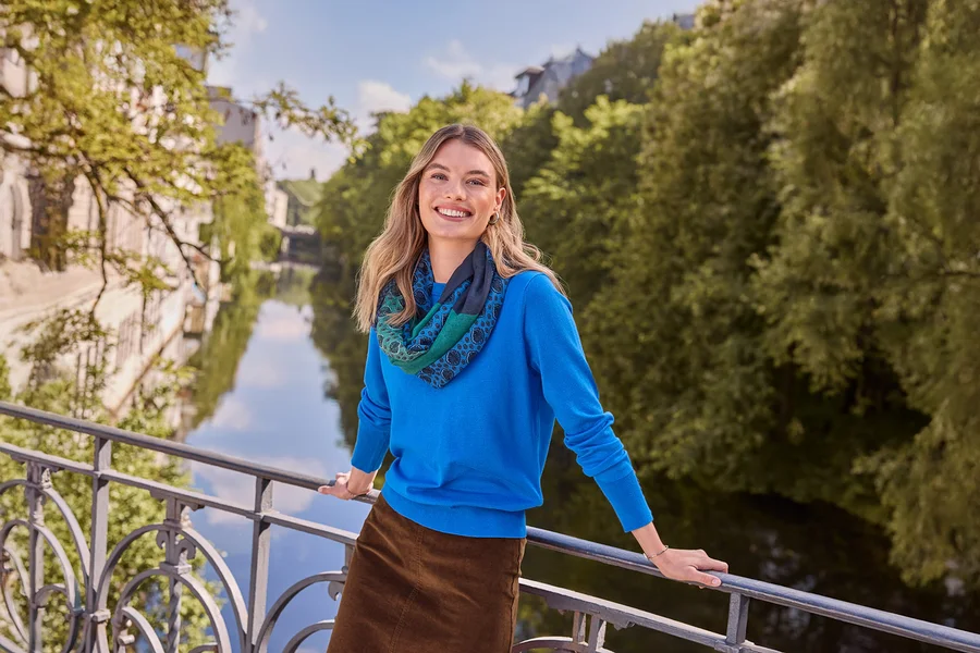 Lächelnde Frau in blauem Oberteil, braunem Rock und gemustertem Schal, trägt Doppel-Creolen aus 925 Silber rhodiniert, lehnt an einer Brücke über einem Fluss.