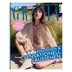 Magazincover für GRAZIA mit einer Frau mit dunklem Haar, die in einem hellen, fließenden V-Ausschnitt-Kleid und geschnürten Stiefeletten hockt, geschmückt mit einer verzierten rosa-weißen Halskette, vor einem grasbewachsenen Hintergrund. Text: „HIGH JEWELLERY ISSUE“ und „ANZIEHEN & SENSATIONELL AUSSEHEN“.