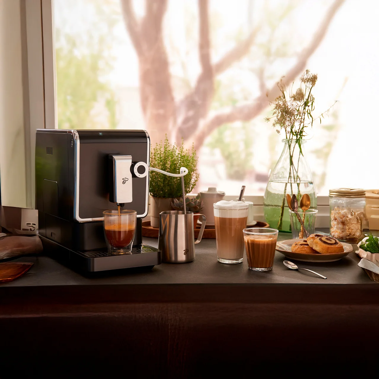 Kaffeemaschine bereitet Kaffee in einem Glas zu. Auf dem Tisch stehen ein Milchaufschäumer, Latte Macchiato und Gebäck.
