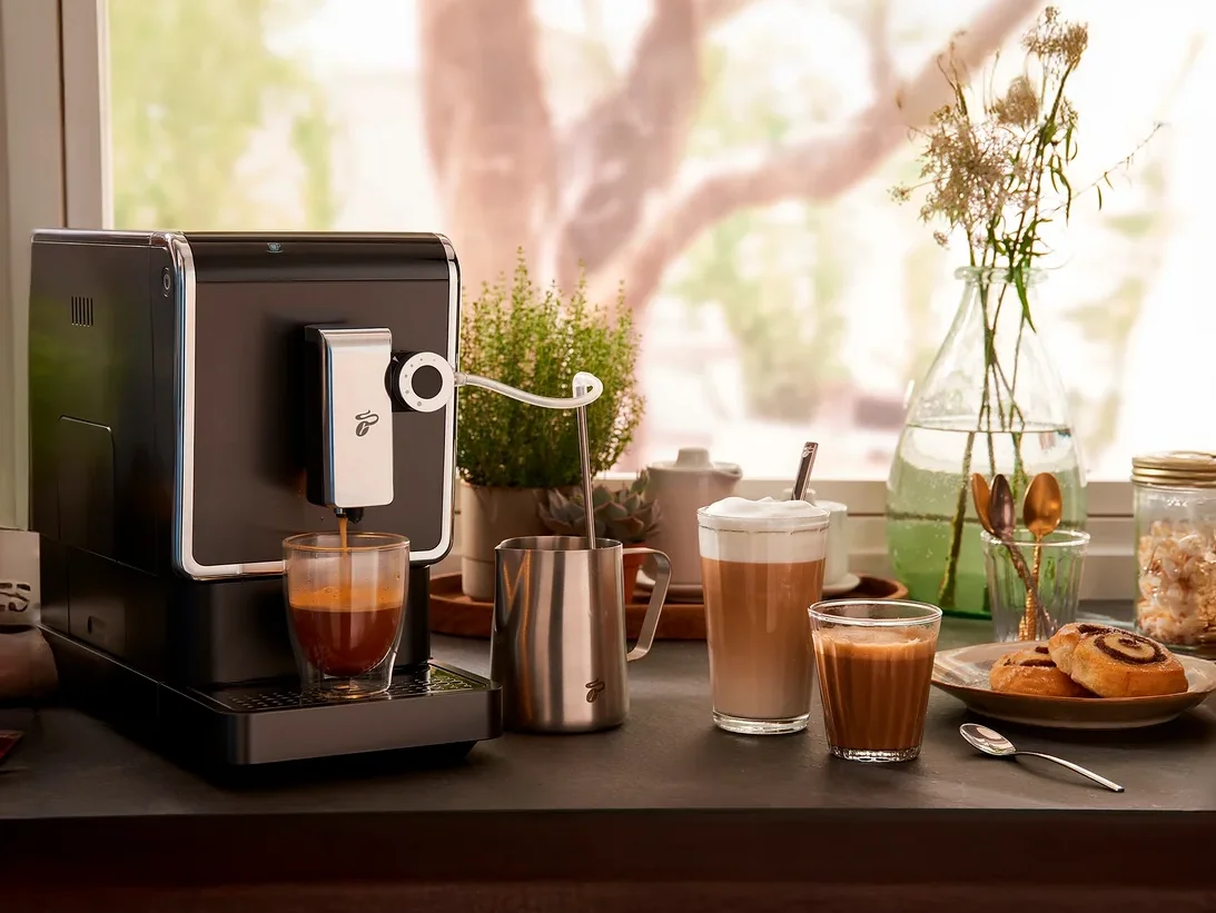 Kaffeemaschine bereitet Kaffee in einem Glas zu. Auf dem Tisch stehen ein Milchaufschäumer, Latte Macchiato und Gebäck.