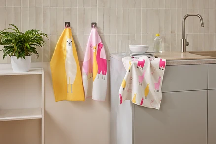 Drei Geschirrtücher mit Lama-Motiven hängen in einer Küche neben einem Waschbecken und einem Regal.