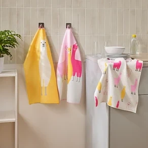 Drei Geschirrtücher mit Lama-Motiven hängen in einer Küche neben einem Waschbecken und einem Regal.