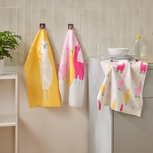 Drei Geschirrtücher mit Lama-Motiven hängen in einer Küche neben einem Waschbecken und einem Regal.
