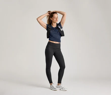 Frau posiert in Outdoor-Leggings, marineblauem Shirt und Weste vor hellem Hintergrund.