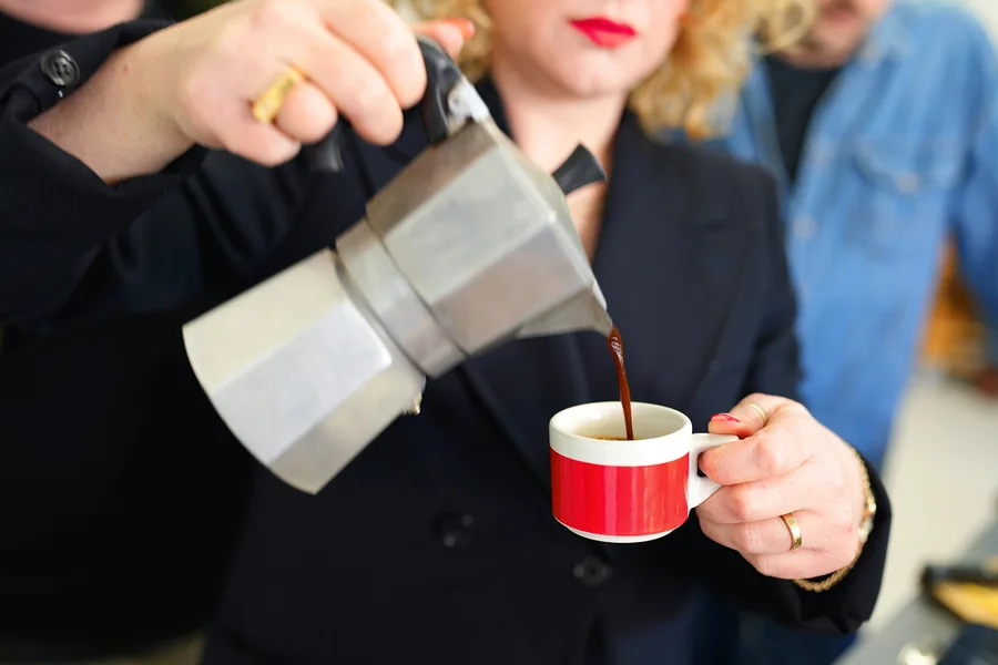 Frau gießt Kaffee aus einer Aluminium-Mokakanne in eine rot-weiße Tasse.