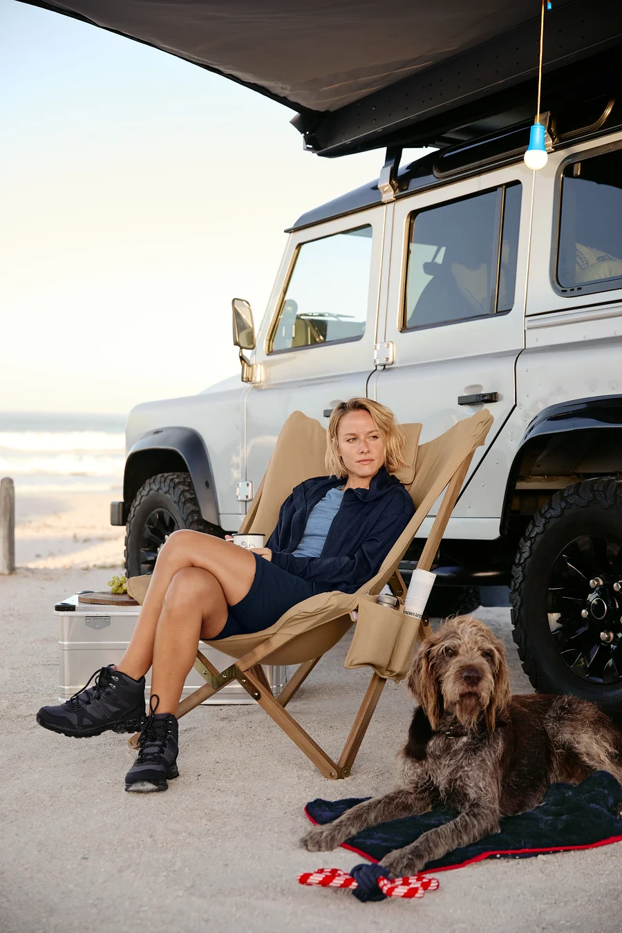Eine Frau sitzt in einem Relaxsessel mit einem Becher. Neben ihr liegt ein Hund auf einer Hunde-Kuscheldecke mit einem Hunde-Spielzeug, Tau. Im Hintergrund sind ein Auto, eine LED-Camping-Lampe, eine 2-in-1-Funktionshose und eine Kapuzenjacke.