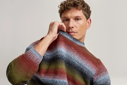 Mann mit lockigem Haar, der den Kragen seines blau, rot, grün und braun gestreiften Pullovers hält und in die Kamera blickt.