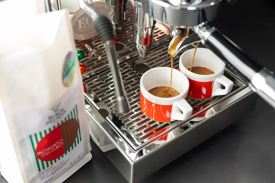 Kaffee läuft in zwei rote Tassen aus einer Kaffeemaschine neben einer Packung Black Delight gemahlenem Kaffee.