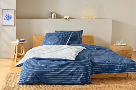 Bett mit blau-weiß gestreifter Bettwäsche und Knotenmuster. Auf dem Nachttisch stehen Bücher und eine Lampe in Schiffsform.