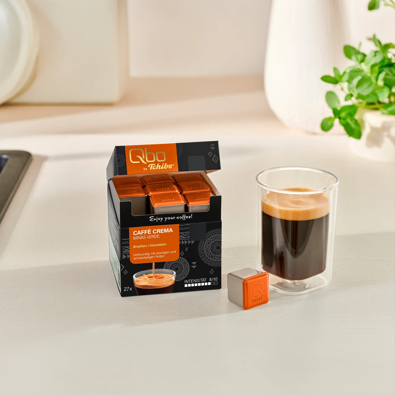 Offene Packung Qbo by Tchibo Caffè Crema Minas Verde Kaffeekapseln mit 27 orangefarbenen Kapseln, daneben ein Glas mit zubereitetem Kaffee und eine einzelne Kapsel auf einer Arbeitsplatte.
