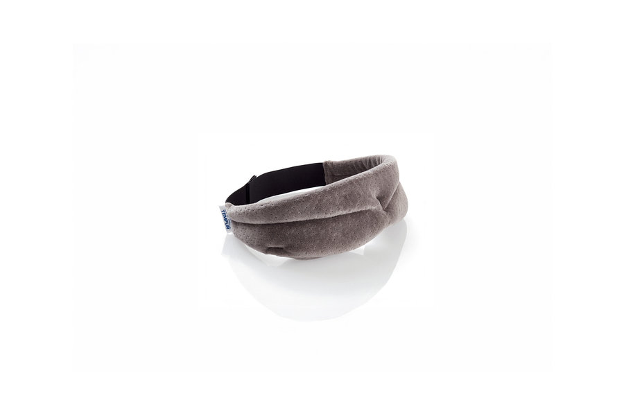 Eine graue, konturierte Schlafmaske mit einem schwarzen Gummiband und einem kleinen weißen „TEMPUR“-Etikett, auf einer weißen Oberfläche mit Reflexion liegend.