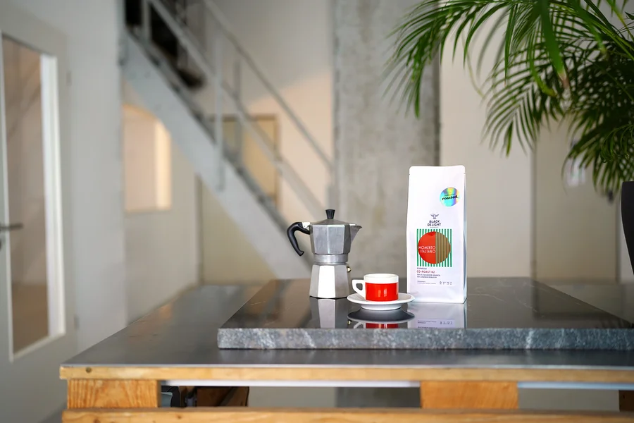Auf einem Tisch stehen eine Kaffeemaschine, eine Tasse und eine Packung Black Delight x roasted. - Momento Italiano Co-Roast #2 Espresso.