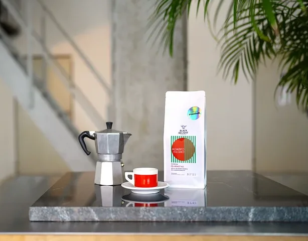 Auf einem Tisch stehen eine Kaffeemaschine, eine Tasse und eine Packung Black Delight x roasted. - Momento Italiano Co-Roast #2 Espresso.