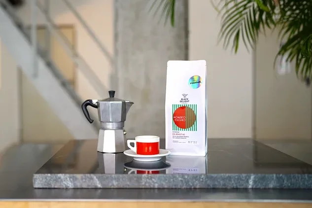 Auf einem Tisch stehen eine Kaffeemaschine, eine Tasse und eine Packung Black Delight x roasted. - Momento Italiano Co-Roast #2 Espresso.