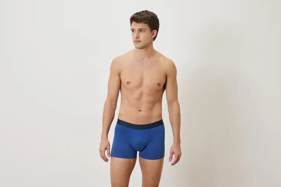 Mann mit freiem Oberkörper und blauen Boxershorts vor einem weißen Hintergrund.
