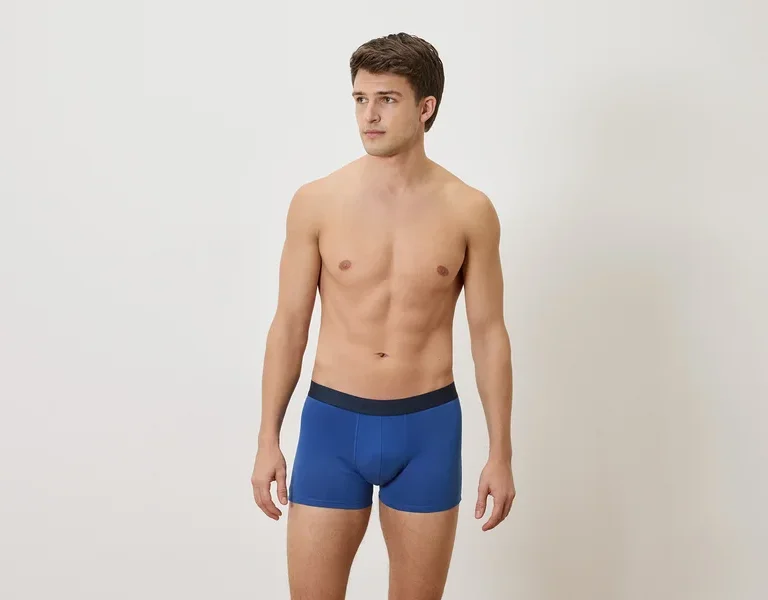 Mann mit freiem Oberkörper und blauen Boxershorts vor einem weißen Hintergrund.