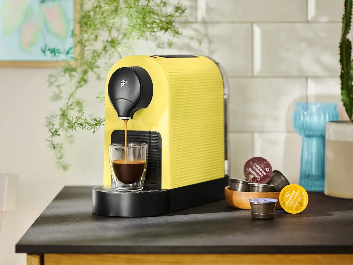 Eine gelbe Kaffeemaschine füllt eine Tasse mit Kaffee, Kapseln liegen daneben.