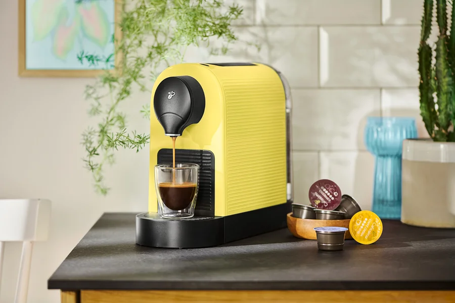 Eine gelbe Kaffeemaschine füllt eine Tasse mit Kaffee, Kapseln liegen daneben.