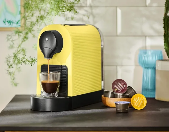Eine gelbe Kaffeemaschine füllt eine Tasse mit Kaffee, Kapseln liegen daneben.