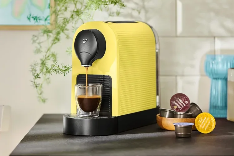 Eine gelbe Kaffeemaschine füllt eine Tasse mit Kaffee, Kapseln liegen daneben.