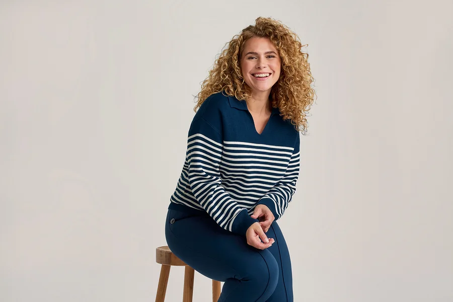 Lachende Frau mit Locken, die einen marineblauen und weiß gestreiften Pullover und marineblaue Hosen trägt, sitzt auf einem Holzschemel.