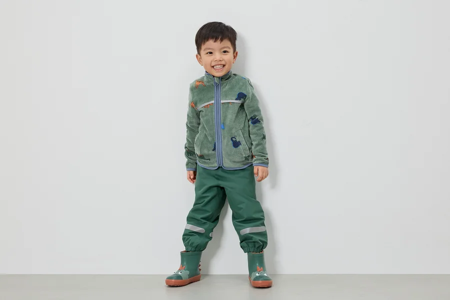 Ein lächelnder Junge steht in grüner Fleecejacke mit Dinosauriern, grüner Hose und grünen Gummistiefeln vor einer weißen Wand.