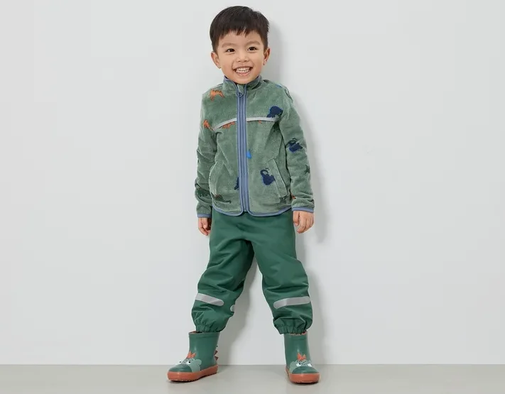 Ein lächelnder Junge steht in grüner Fleecejacke mit Dinosauriern, grüner Hose und grünen Gummistiefeln vor einer weißen Wand.