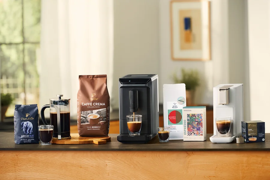 Verschiedene Kaffeeprodukte auf einer Theke: eine blaue Packung Privat Kaffee African Blue, eine French Press mit Kaffee, ein doppelwandiges Caffè Crema Glas, eine braune Packung Caffè Crema Vollmundig, ein grauer Esperto mini Kaffeevollautomat, der Kaffee in ein doppelwandiges Caffè Crema Glas füllt, ein kleines doppelwandiges Caffè Crema Glas, eine weiße Packung Black Delight Espresso, eine bunte sometime CONTEMP 1 Espresso Packung, eine weiße Qbo ICONIC Light Gold Maschine, die Kaffee in ein doppelwandiges Caffè Crema Glas füllt, und eine schwarze Packung Qbo Caffè JABANA YIRGA Kapseln.