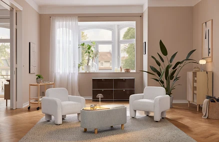 Gemütliches Wohnzimmer mit zwei hellgrauen Sesseln und einem passenden Ottoman auf einem Teppich. Eine beige LED-Akku-Tischleuchte steht auf einem hellen Holzschrank neben einer großen Pflanze. Unter einem Erkerfenster befindet sich ein dunkelbraunes Aufbewahrungsmöbel.