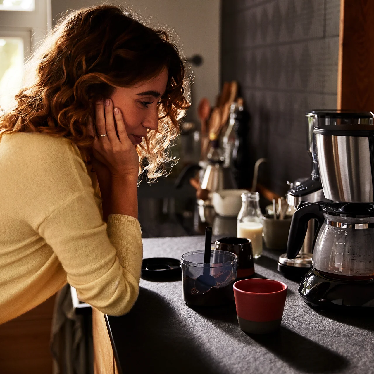 Frau lehnt in der Küche neben einer Kaffeemaschine, einer Tasse und einem Glas.