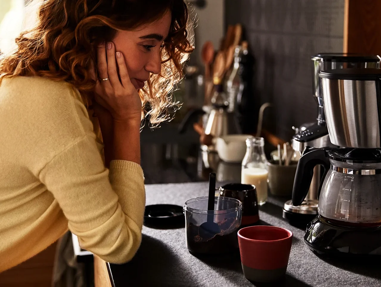 Frau lehnt in der Küche neben einer Kaffeemaschine, einer Tasse und einem Glas.