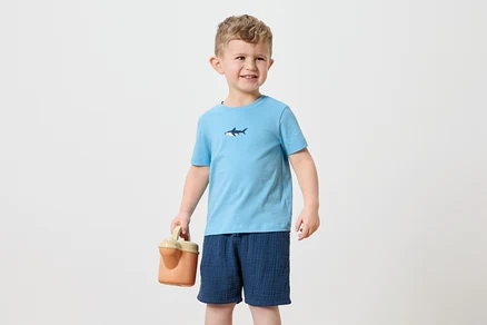 Ein kleiner Junge mit blonden Haaren trägt ein hellblaues T-Shirt mit einem Hai-Aufdruck und marineblaue Shorts. Er hält eine orangefarbene Gießkanne.