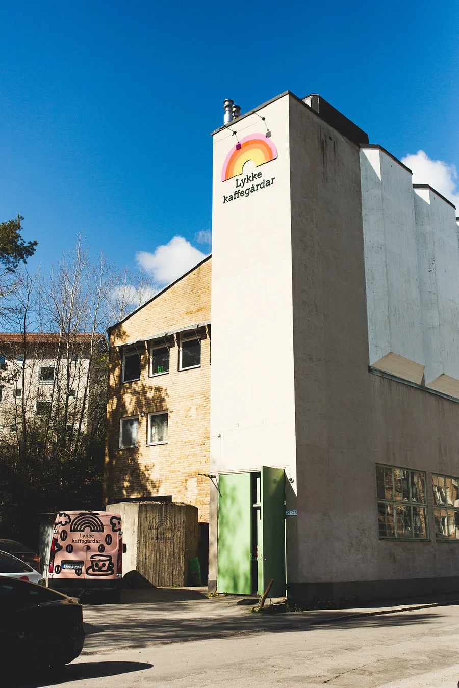 Ein helles Gebäude mit Regenbogen-Logo und „Lykke kaffegårdar“-Schriftzug an der Seite, neben einem Ziegelgebäude. Ein pinker Lieferwagen mit Kaffee-Branding parkt links unter blauem Himmel.