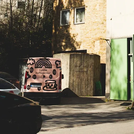 Ein helles Gebäude mit Regenbogen-Logo und „Lykke kaffegårdar“-Schriftzug an der Seite, neben einem Ziegelgebäude. Ein pinker Lieferwagen mit Kaffee-Branding parkt links unter blauem Himmel.