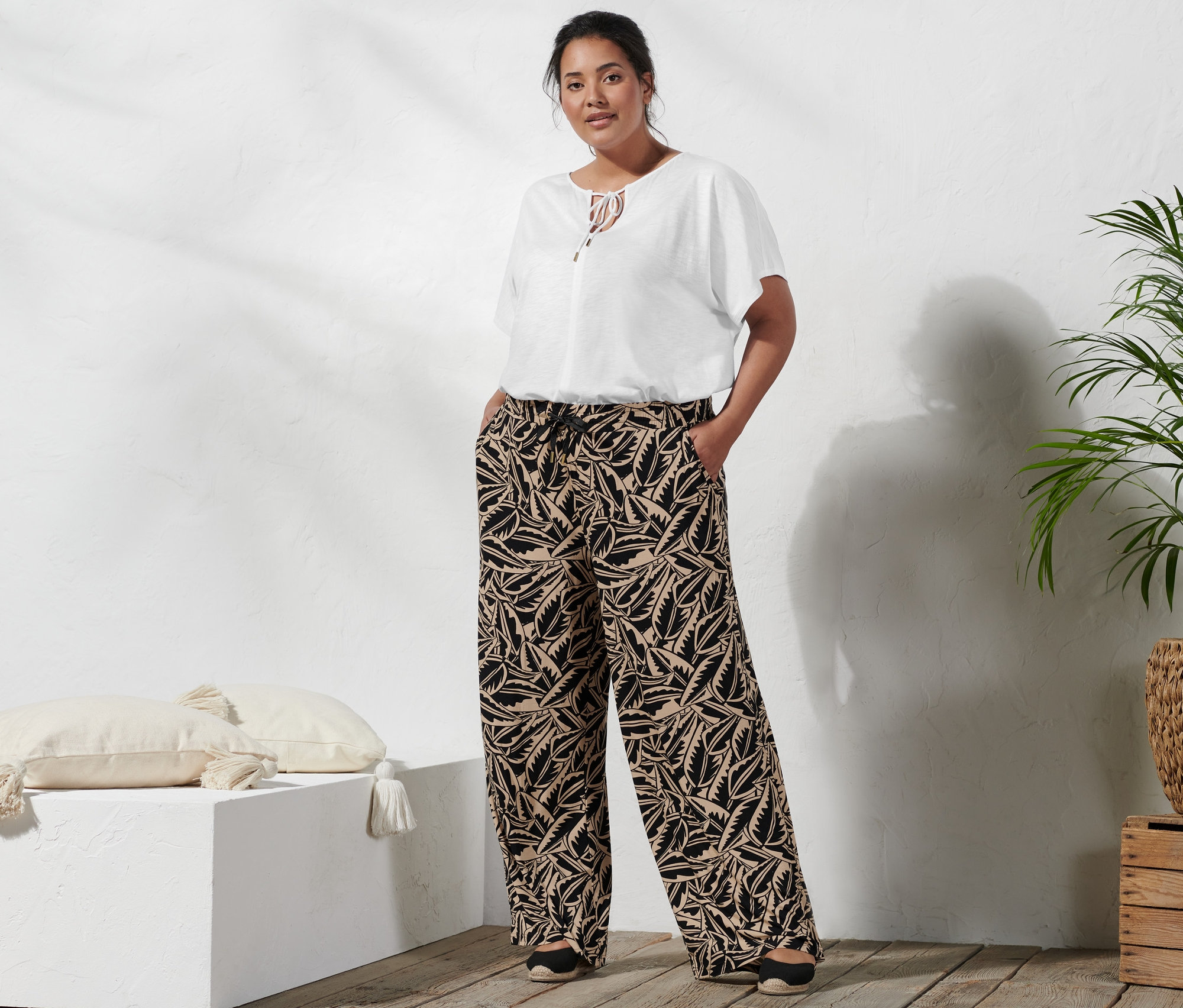 Eine Frau steht mit den Händen in den Taschen einer bedruckten Palazzohose in Schwarz-Beige, dazu trägt sie ein Blusenshirt mit Bindeband und Espadrilles mit Keilabsatz.