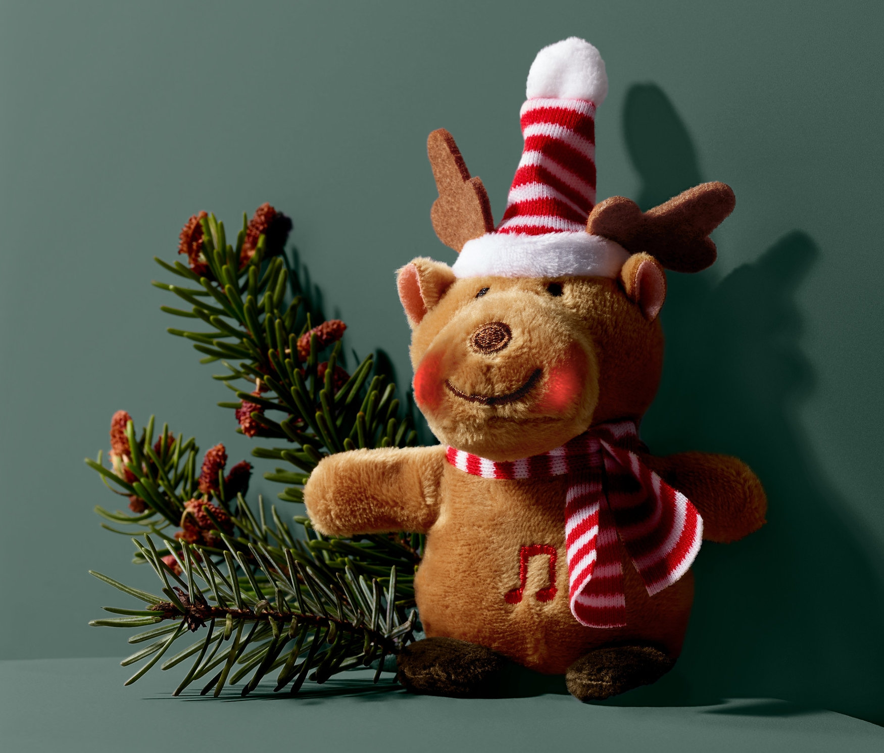 Singendes Rentier mit Schal und Mütze steht neben einem Nadelbaumzweig.