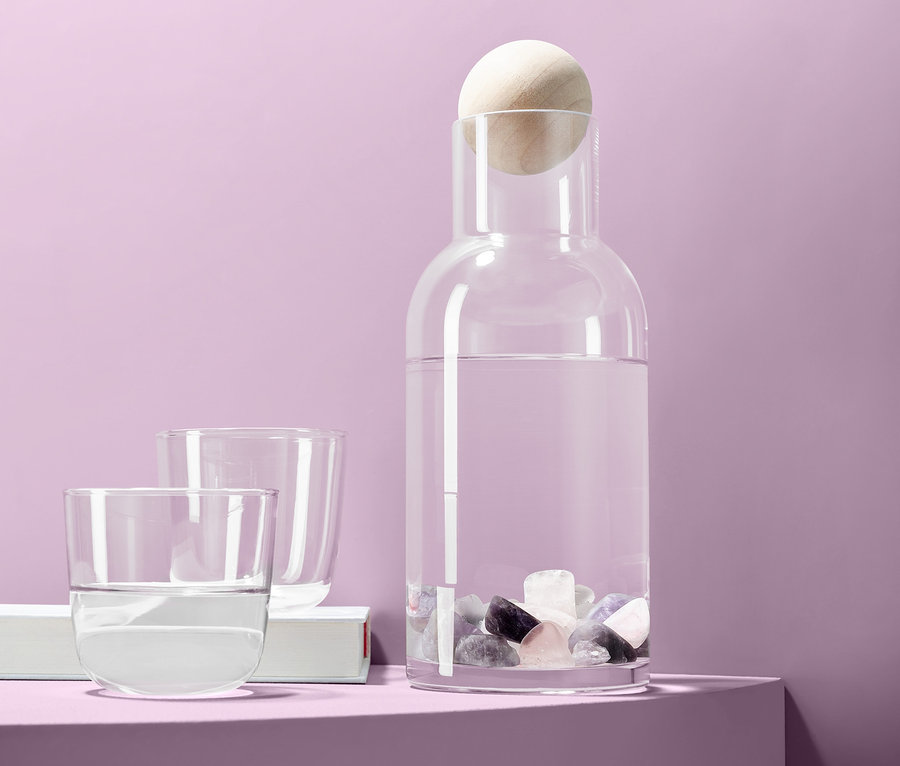 Eine Glaskaraffe mit Wasser und Wassersteine, dazu zwei Gläser, vor einem lila Hintergrund.